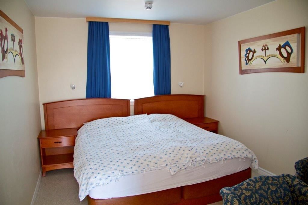A bedroom with a wooden bed, a nightstand, a chair, and a window with blue curtains.