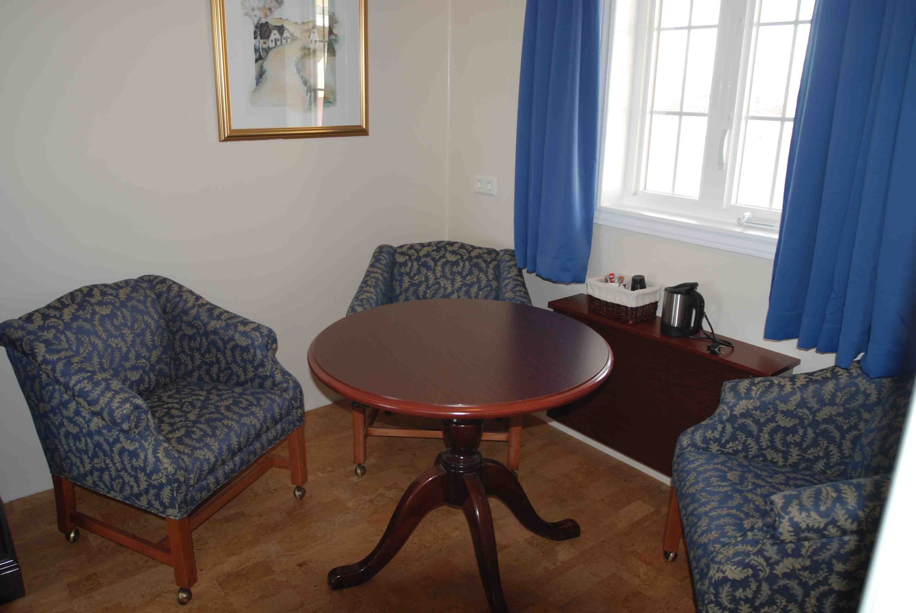 A room with a round wooden table, two patterned armchairs, a small side table with a kettle, and a framed picture on the wall.