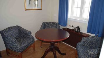A room with a round wooden table, two patterned armchairs, a small side table with a kettle, and a framed picture on the wall.