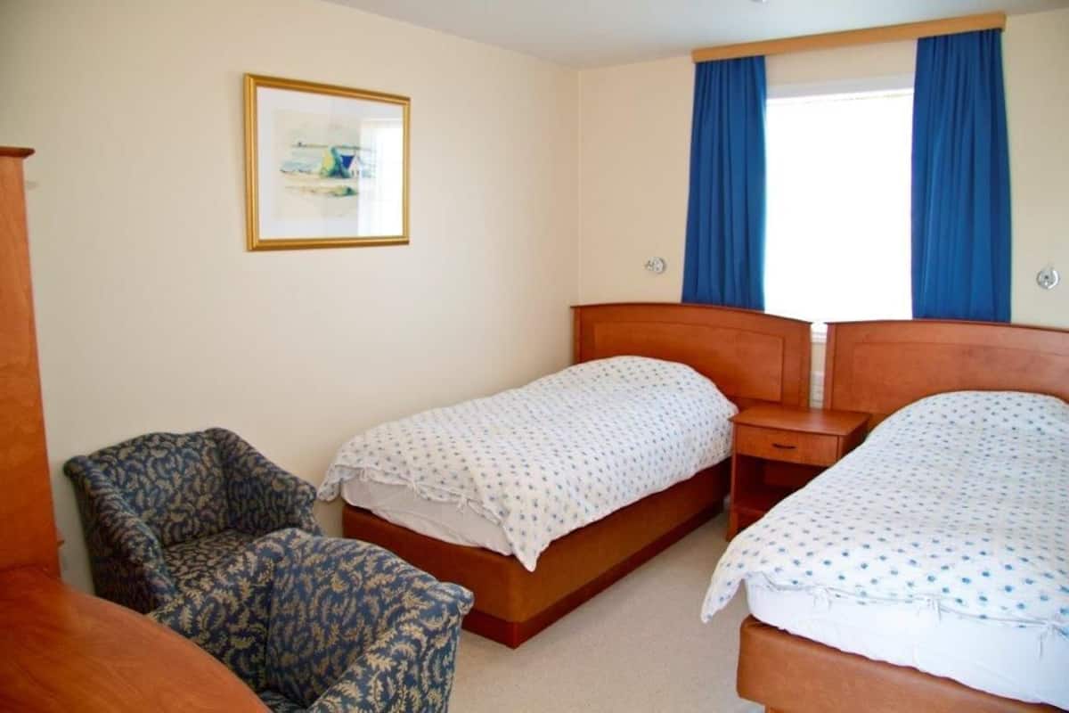 A hotel room with two beds, a chair, a wooden nightstand, a framed picture on the wall, and blue curtains on the window.