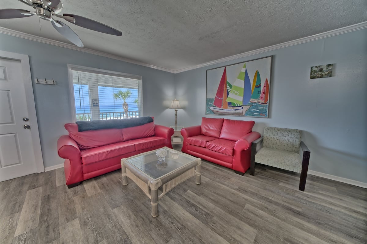 A living room with two red leather sofas, a glass coffee table, a ceiling fan, a window with blinds, and a sailboat painting on the wall.