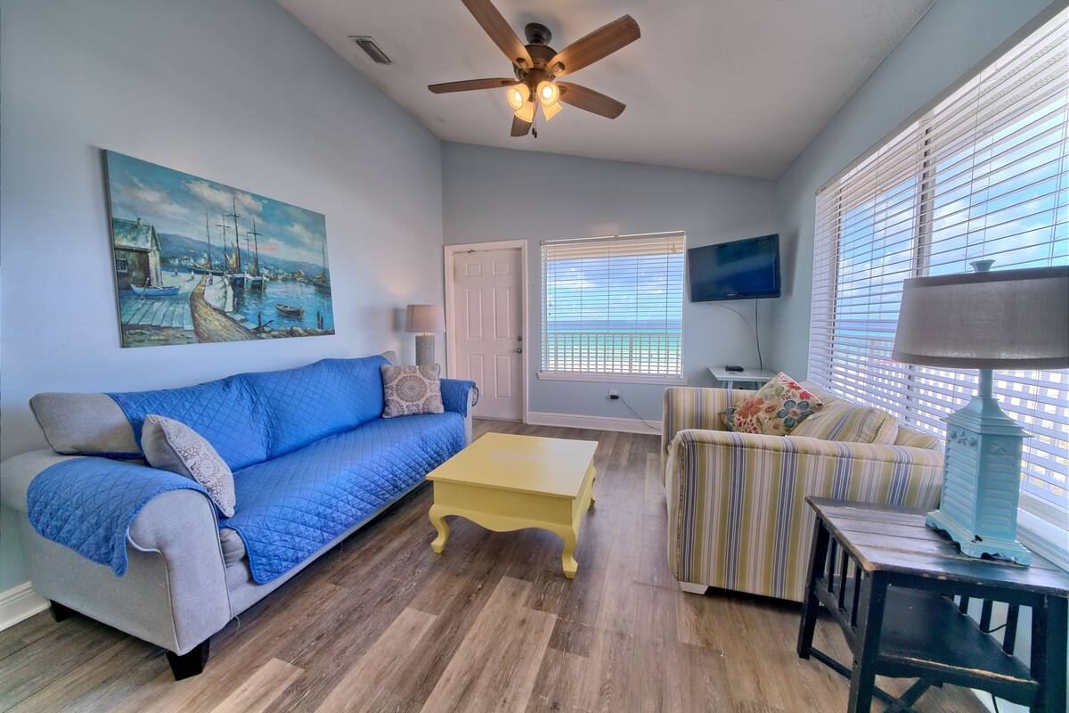 A living room with a blue sofa, a yellow coffee table, a striped armchair, a ceiling fan, and a painting on the wall.
