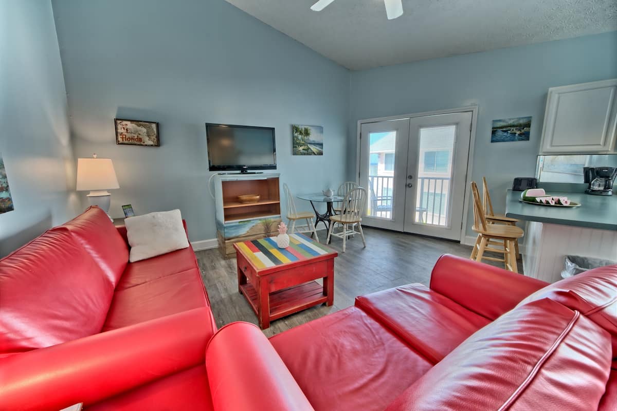 A living room with red leather sofas, a TV, a small table, and a balcony with a view.