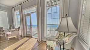 Een ruime kamer met een houten vloer, een groot raam met uitzicht op zee, een chaise longue met een zwanendecoratie, en een lamp op een bijzettafeltje.