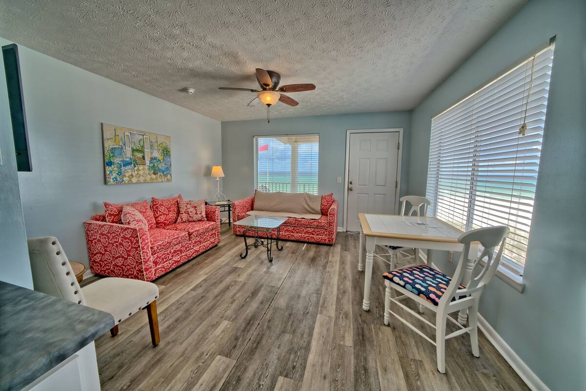 A living room with a red patterned sofa, a wooden coffee table, a ceiling fan, a dining table with chairs, and a window with blinds.