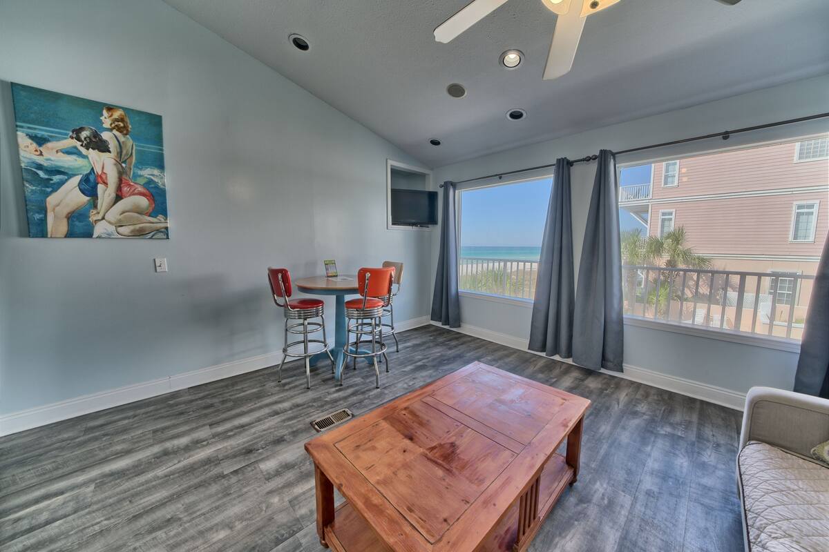 A room with a wooden table, two red chairs, a painting, a TV, and a large window with a view of the ocean.