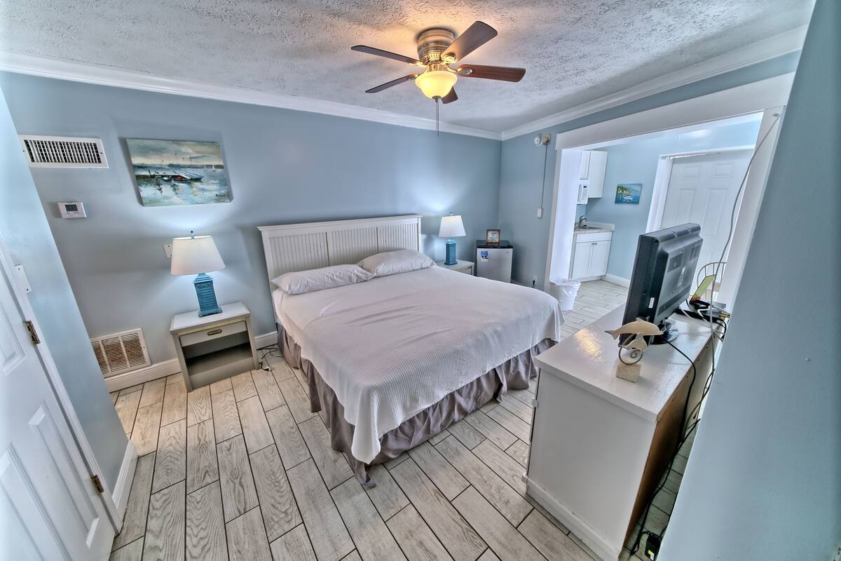 A bedroom with a bed, bedside tables, a ceiling fan, a TV, and a view of the adjacent bathroom.