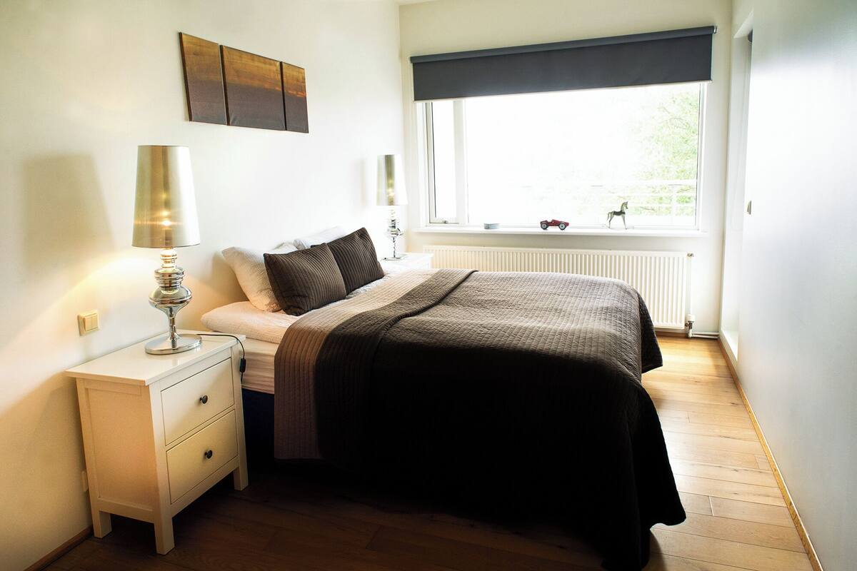A bedroom with a bed, bedside table, lamp, window with blinds, radiator, and a toy on the sill.