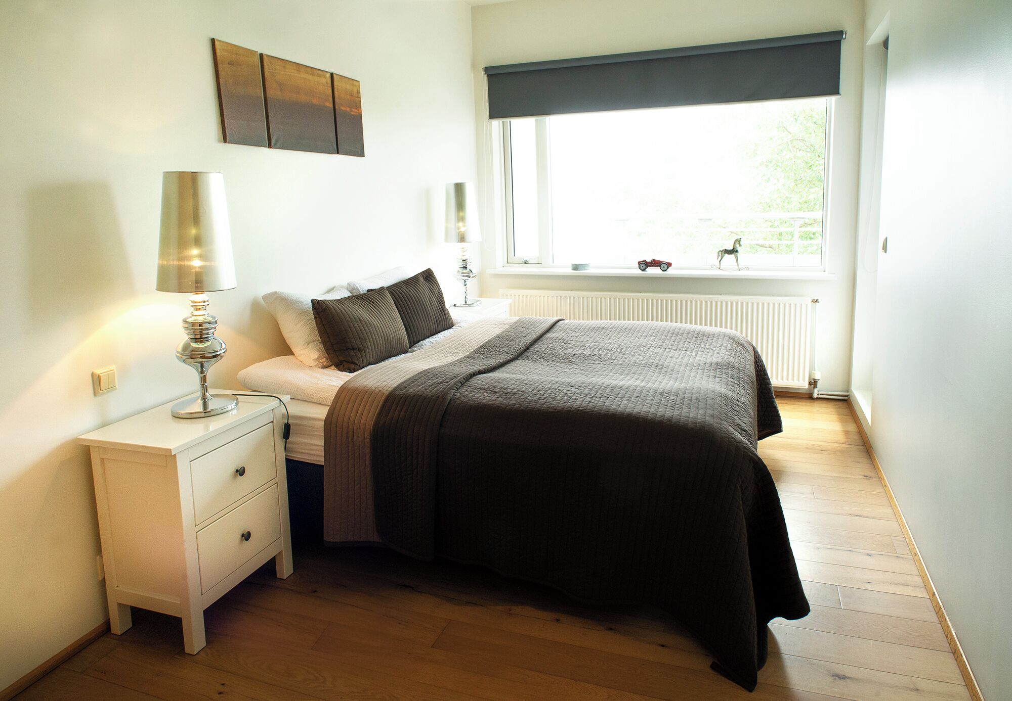 Un dormitorio con cama, mesita de noche, lámpara, ventana con vistas y un radiador.
