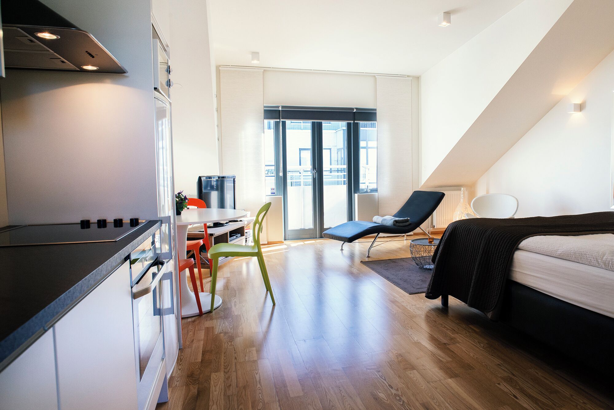 A modern, well-lit room with a kitchenette, a dining area, a lounge chair, and a bed.