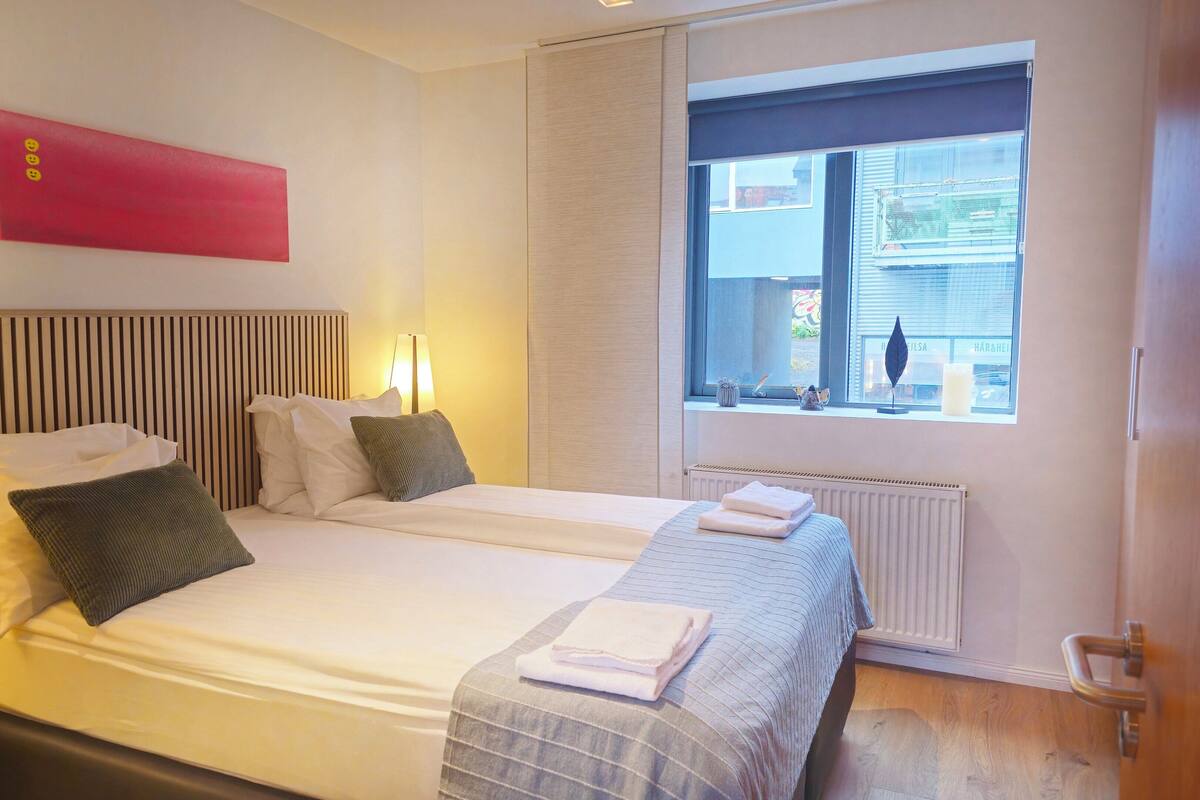 A double bed with two pillows, a bedside table with a lamp, a window with a view of the outside, and a red abstract art piece on the wall.