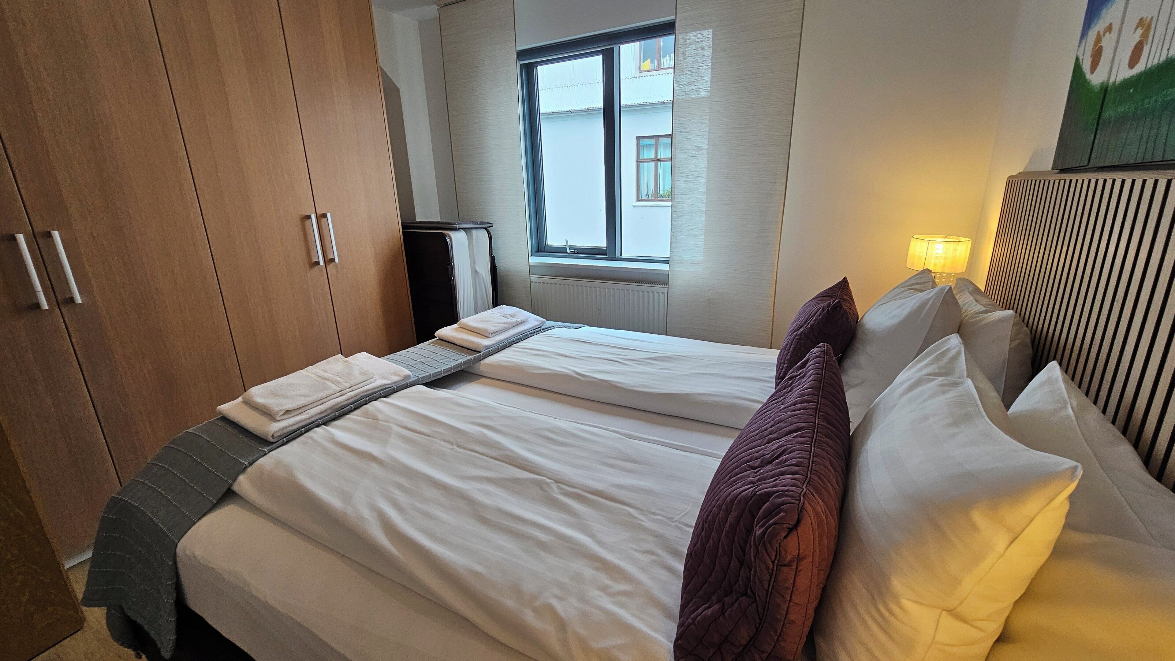 A hotel room with a bed, a window, a radiator, and a lamp.