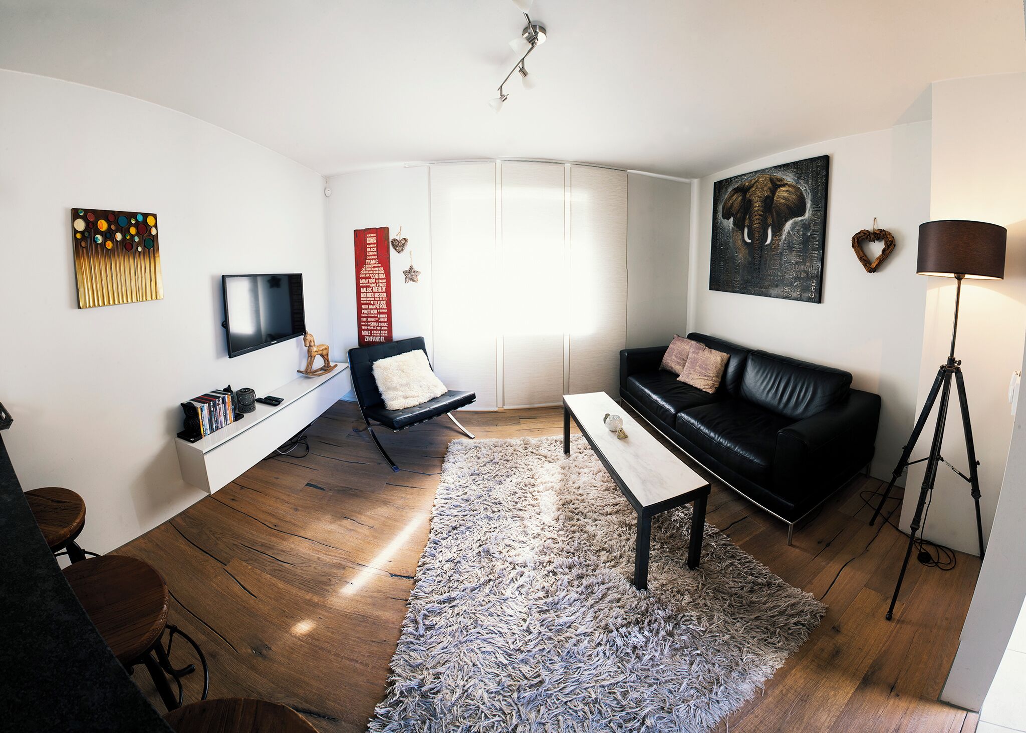 Una sala moderna con un sofá de cuero negro, una mesa de centro blanca, una silla negra, un aparador blanco, un televisor montado en la pared y una lámpara de pie.