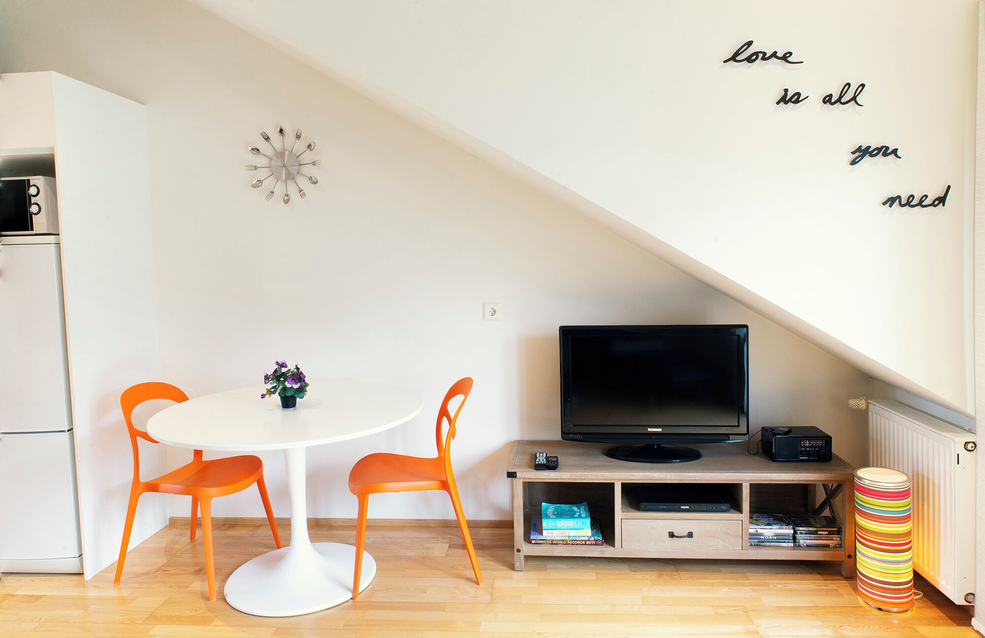 A modern living space with a white dining table, orange chairs, a flat-screen TV, a small plant, and a motivational wall quote.