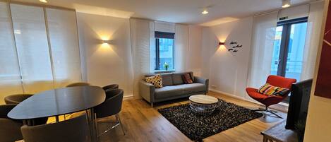 A modern living room with a sofa, a dining table with chairs, a red chair, a TV, and a black rug.