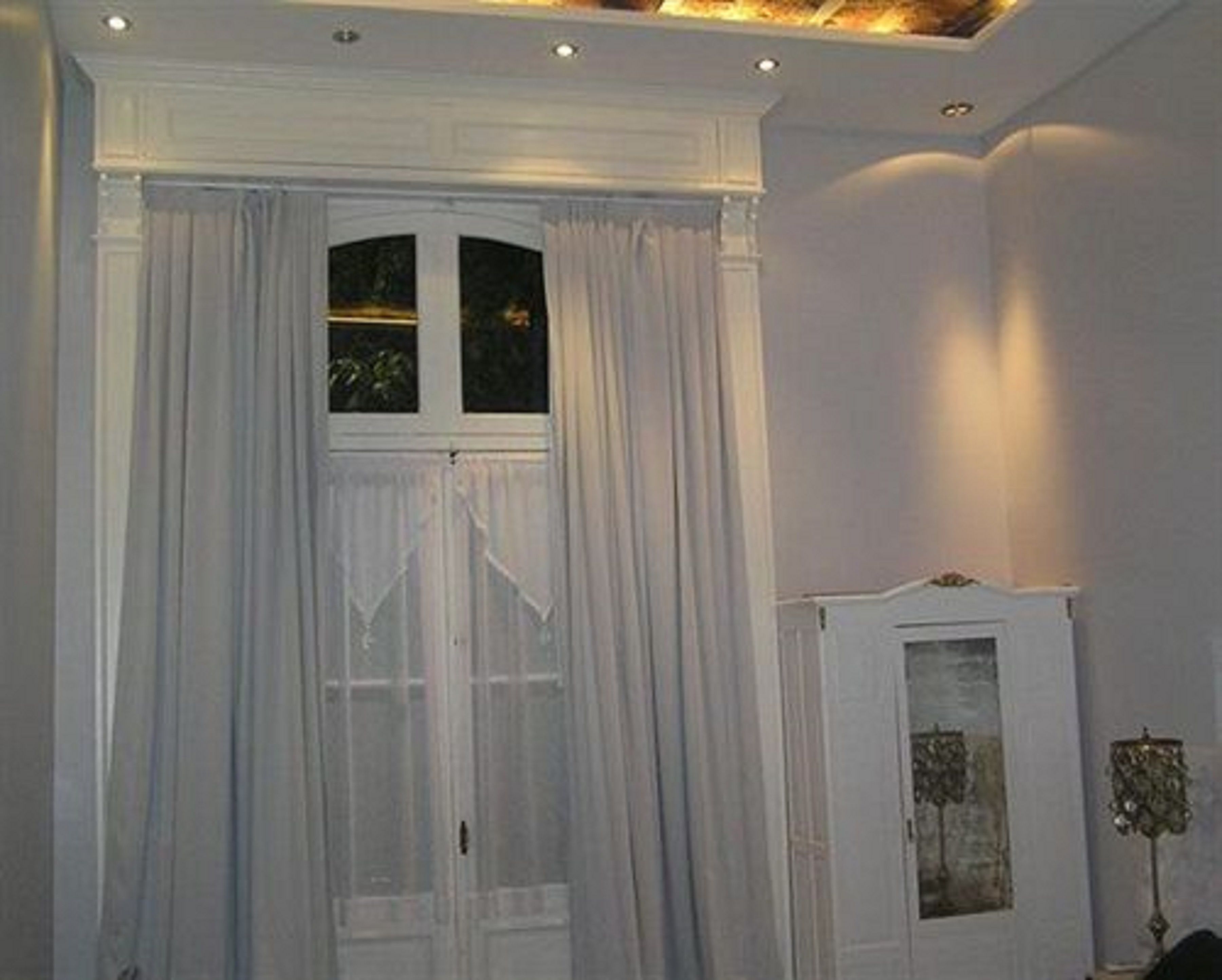 A room with a large window covered by sheer white curtains, a white cabinet with a glass door, and recessed ceiling lighting.