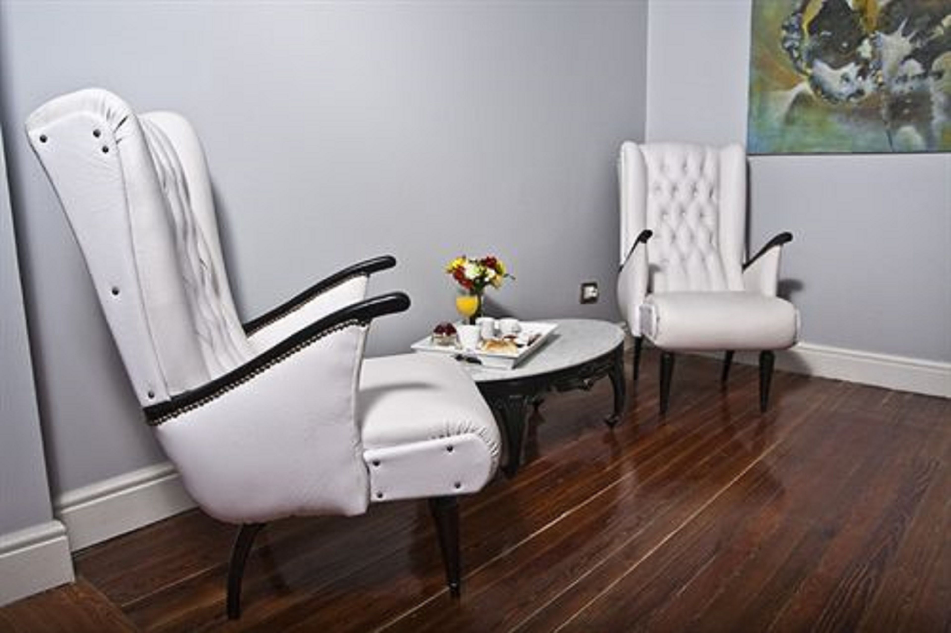 A room with two white upholstered chairs, a small round table with a vase of flowers, and a painting on the wall.