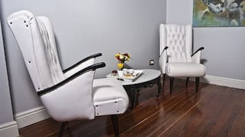 A room with two white upholstered chairs, a small round table with a vase of flowers, and a painting on the wall.