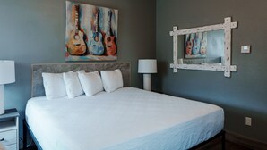 A hotel room with a large bed, a headboard, a painting of guitars, a wall-mounted mirror, and a nightstand with a lamp.