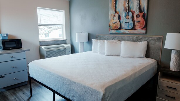 A bedroom with a bed, bedside lamps, a window, a microwave, and a painting of guitars.