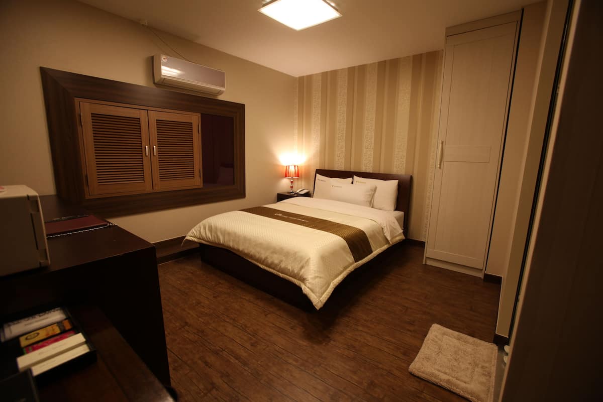 Una habitación de hotel con una cama de madera, un escritorio con libros, una lámpara y un armario.
