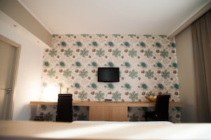 Quarto de hotel com uma cama, televisão na parede, secretária de madeira com um alto-falante e parede decorativa com padrão floral.