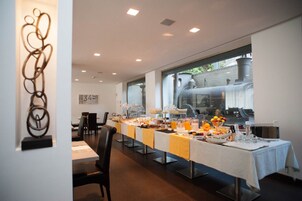 Een lange eettafel met ontbijtartikelen, een kunstwerk en uitzicht op een gebouw door een groot raam.