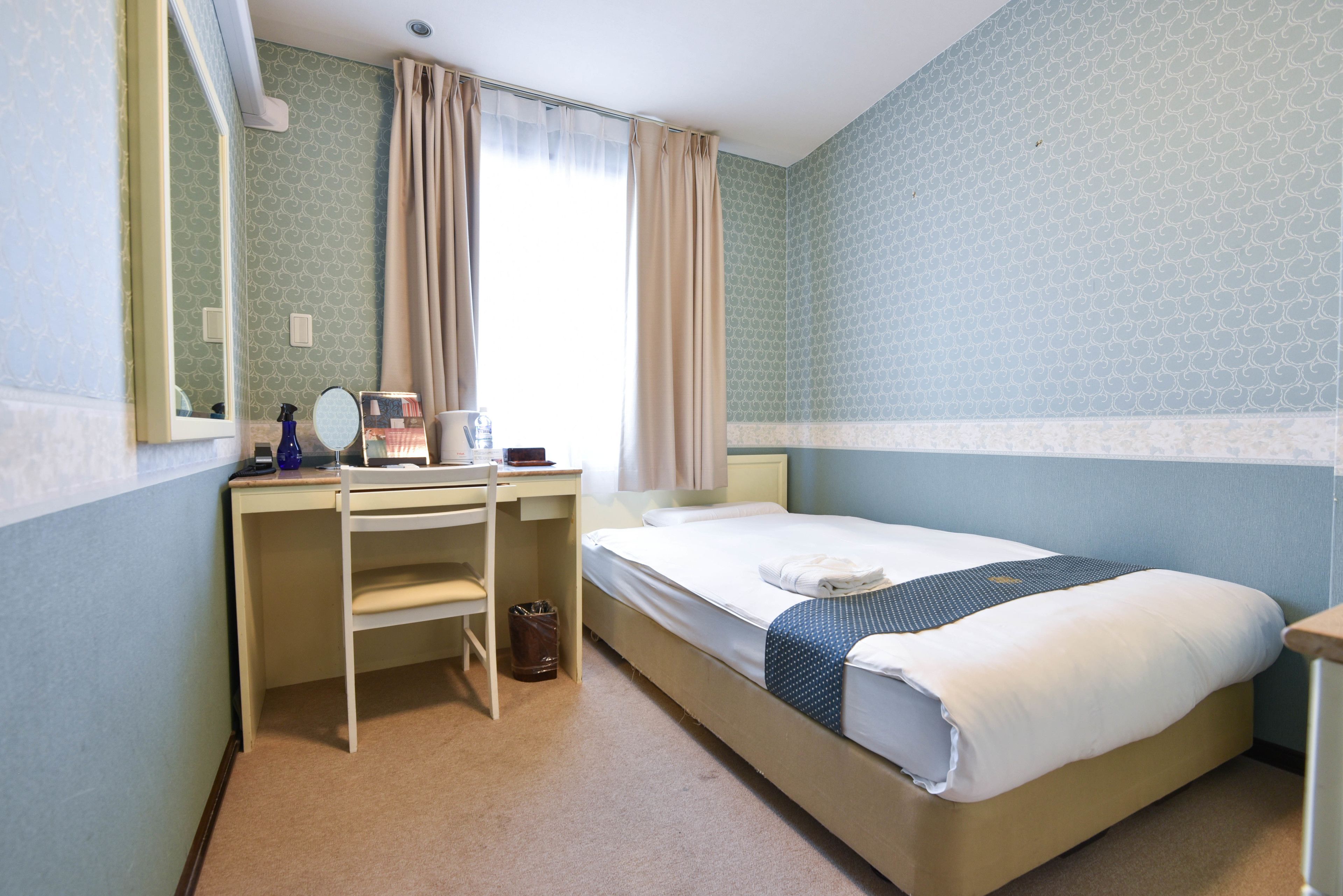 Une petite chambre à coucher, bien rangée, avec un lit simple, un bureau avec une chaise, un miroir et une fenêtre avec des rideaux.