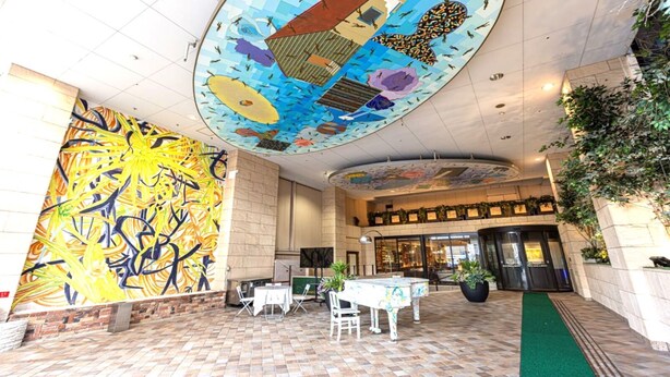 Un vestíbulo amplio con techo alto, un mural grande en la pared de la izquierda y un área de restaurante con mesas al aire libre.