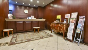 Un área de recepción con un mostrador de madera, dos taburetes, un reloj y una exhibición de folletos y mapas.