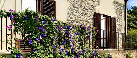 A building with stone walls, wooden shutters, and a balcony with purple flowers.