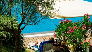 A poolside area with a white umbrella, a person relaxing on a lounge chair, and a tree in the foreground.