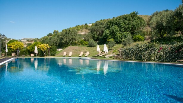 A clear blue swimming pool with lounge chairs and umbrellas, surrounded by greenery and hills.