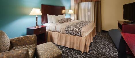 A hotel room with a bed, two bedside tables with lamps, a chair, a desk with a television, and a patterned carpet.