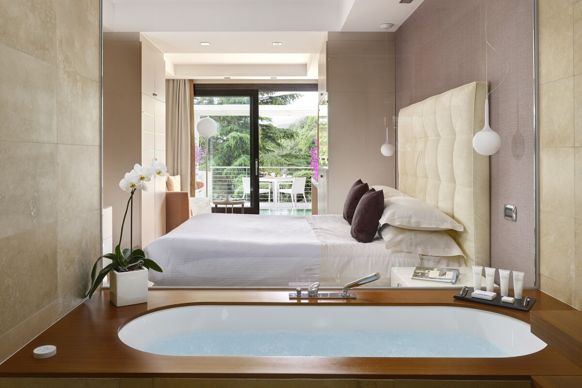 A modern hotel room with a large bathtub, a bed with white linens, and a balcony with a view of greenery.