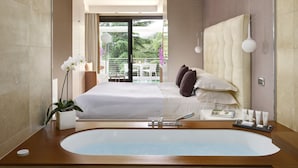 Habitación de hotel moderna con una bañera grande, cama con ropa blanca y balcón con vistas a zonas verdes.