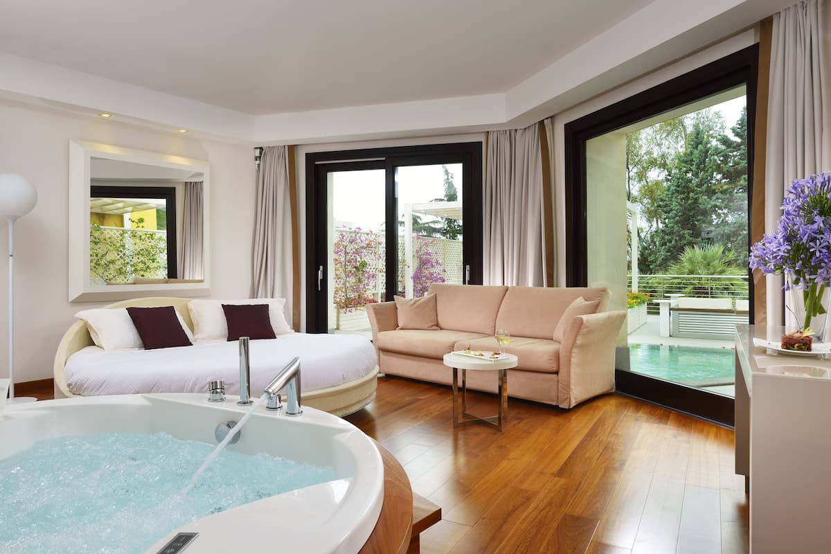 Ein modernes Hotelzimmer mit einem großen Bett, einer Couch, einem kleinen Tisch, einer Badewanne und einem Balkon mit Blick auf einen Pool.