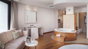 Una habitación de hotel moderna con sofá, una mesita con una copa de vino, un banco de madera redondo, un espejo grande, un baño con bañera y un inodoro.