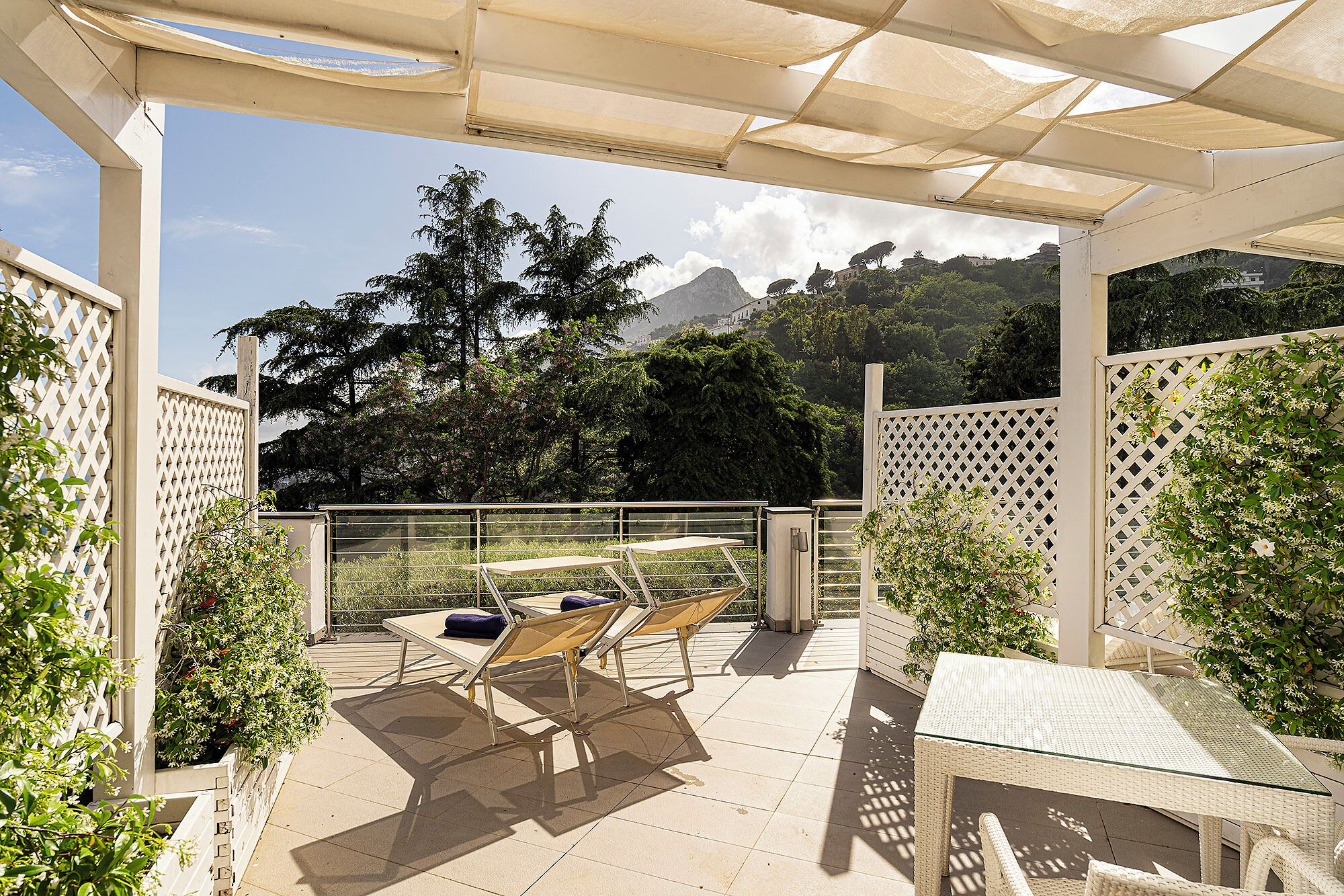 An outdoor patio with a table and two lounge chairs, surrounded by a white lattice fence, overlooking a mountainous landscape.