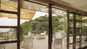 Una terraza moderna al aire libre con mobiliario de mimbre blanco, barandilla de vidrio y vistas a una colina.