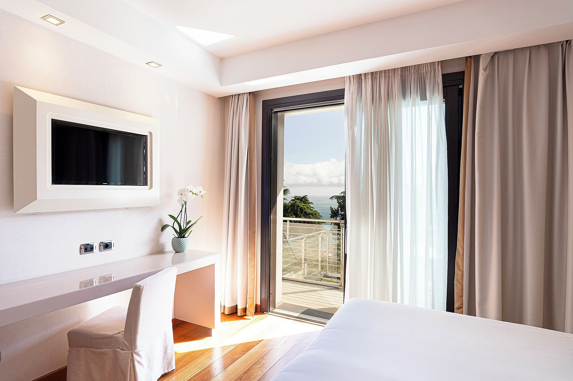 A modern hotel room with a bed, a desk with a TV, a potted plant, and a balcony with curtains.