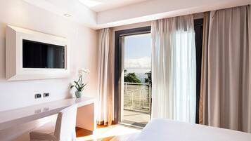 A modern hotel room with a bed, a desk with a TV, a potted plant, and a balcony with curtains.