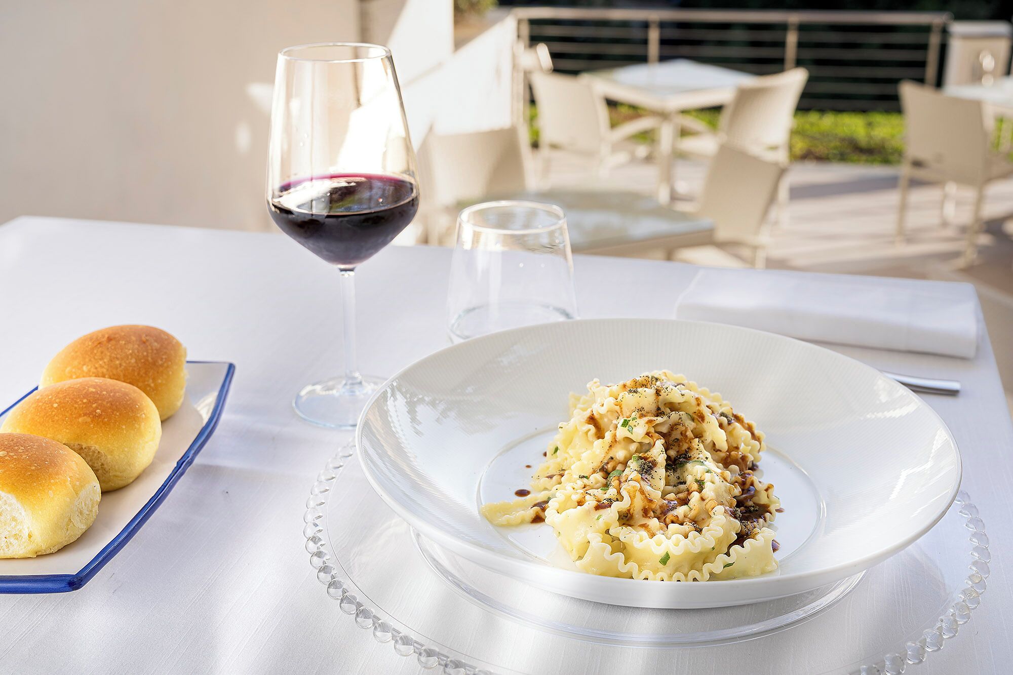 Una mesa de comedor con un plato de pasta, una copa de vino tinto y panecillos.