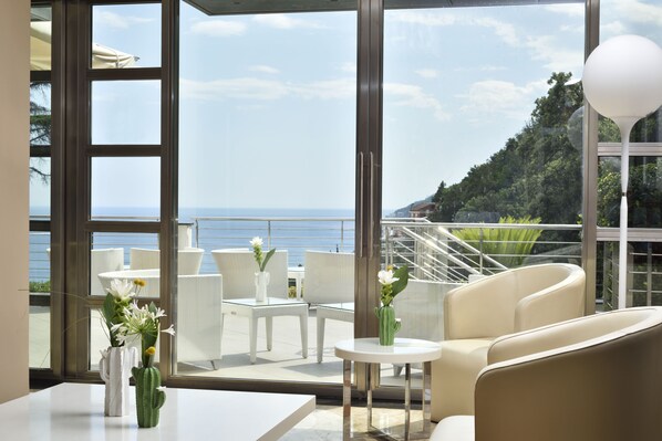 Un espace extérieur moderne, aménagé avec des meubles blancs, une petite table agrémentée d’un vase de fleurs, et offrant une vue sur la mer et la verdure.