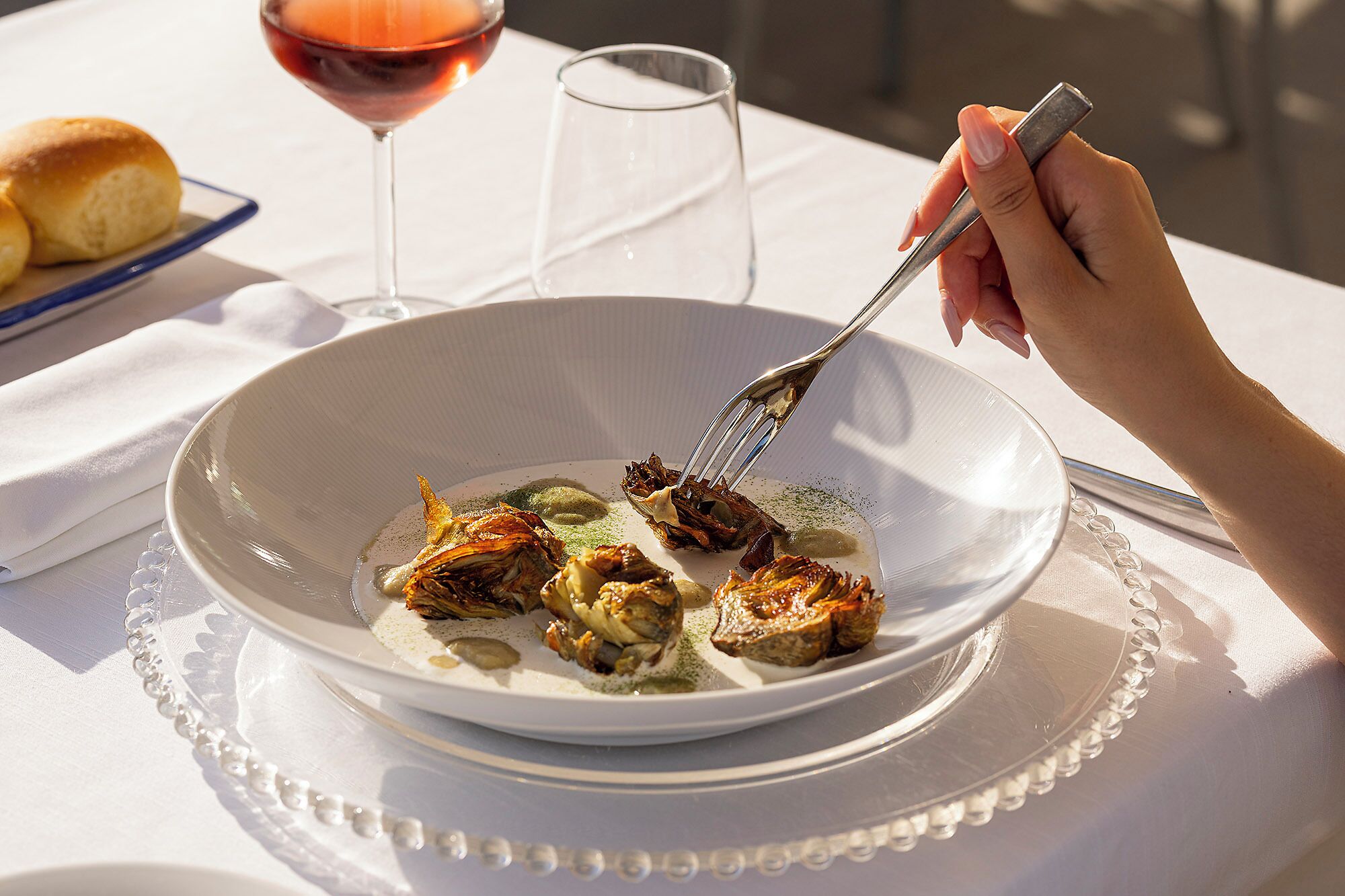 A dining table with a white tablecloth, a plate of food, a glass of red wine, and a bread roll.