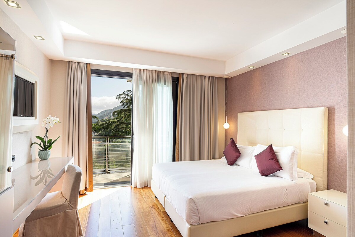 A hotel room with a bed, bedside tables, a desk, and a view of the outdoors.