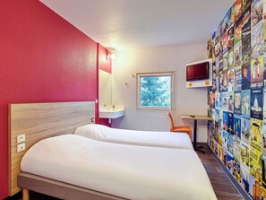 A hotel room with a bed, a desk, a TV, and a wall decorated with travel posters.