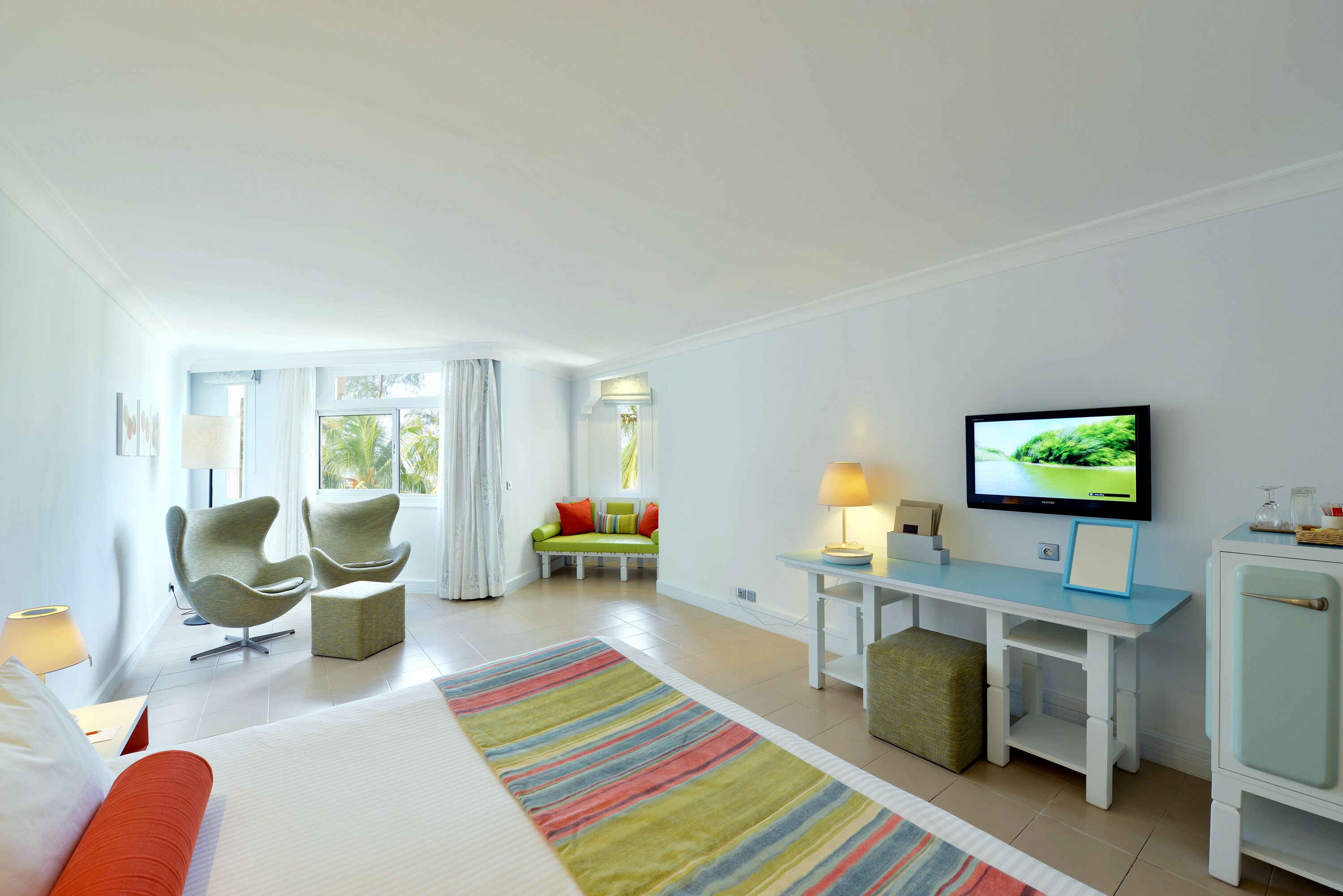 A modern hotel room with a bed, a TV, a desk, and a seating area with chairs and a sofa.