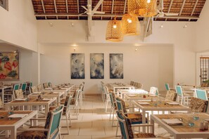 A spacious dining area with white tables and chairs, featuring a high ceiling with exposed beams and hanging wicker lamps.