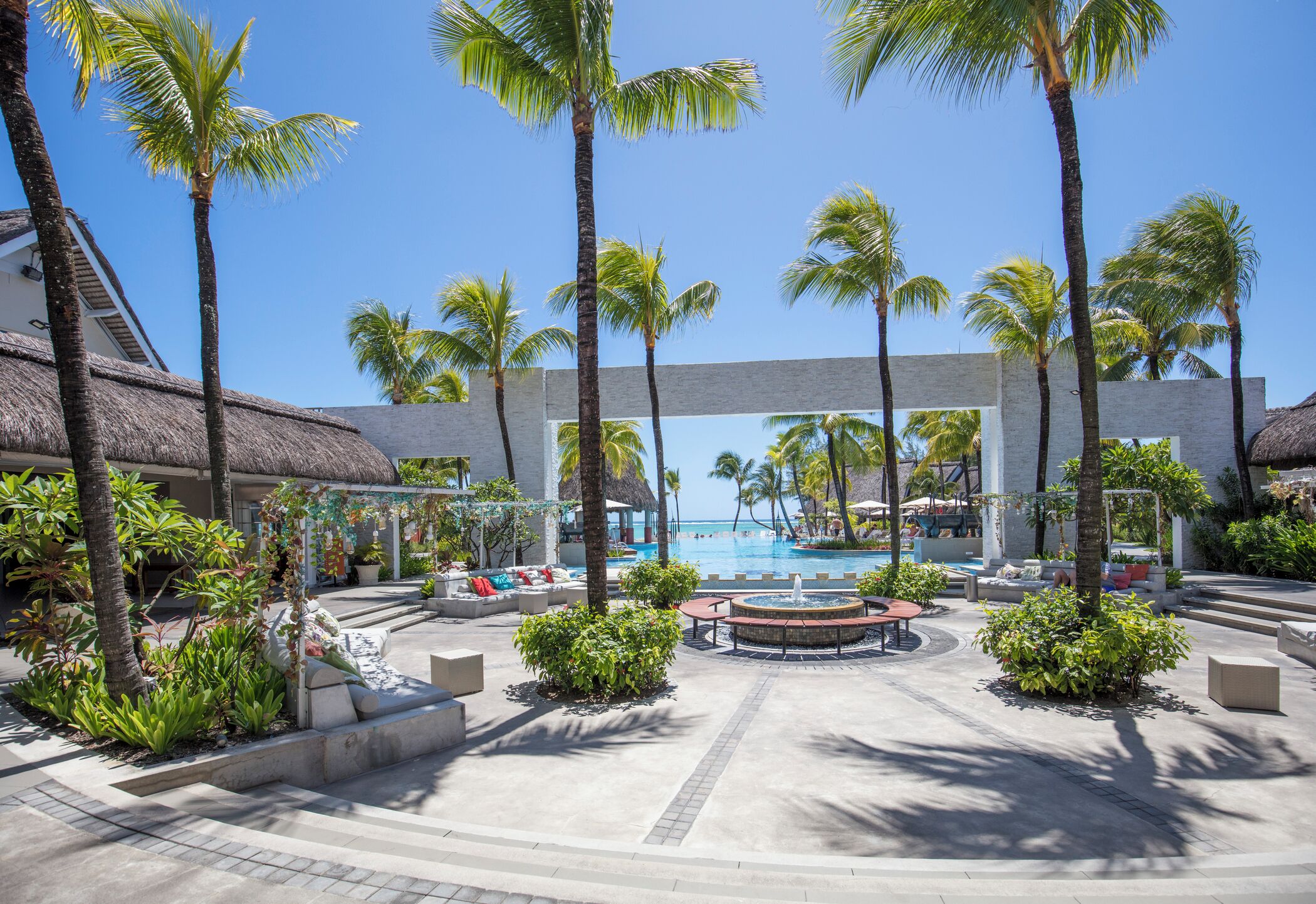 Poolbereich eines Resorts mit Palmen, einem Gebäude mit Reetdach und blauem Himmel.
